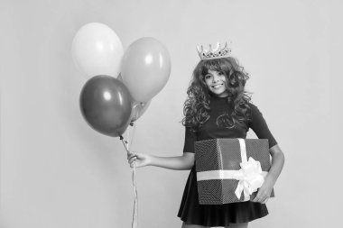 smiling teen child in crown with present box and party balloon on yellow background.