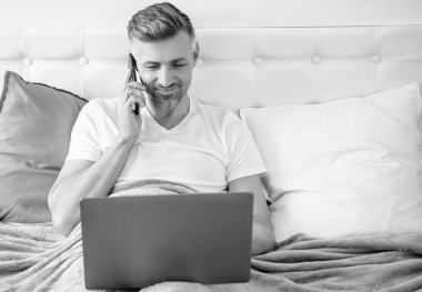 Olgun bir adam telefonda konuşuyor ve laptopla yatakta çalışıyor..