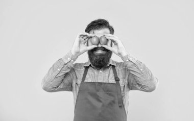 Happy guy in apron holding fresh limes on eyes yellow background, fruit seller.