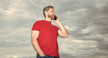 caucasian man has phone talk outdoor. man has phone talk wear red tshirt. photo of man has phone talk. man has phone talk on sky background.