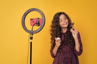 Uzun kıvırcık saçlı, gülümseyen çocuk blogcu blog tutuş pudrası fırçası videosu yapıyor.