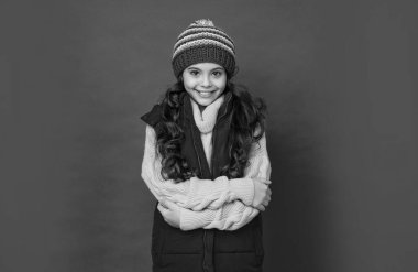 Şapkalı, kıvırcık saçlı mutlu çocuk ellerini çaprazladı. Kadın manken. Yelekli genç kız. Kalın giysiler giyen çocuk portresi. Olumlu duygularını ifade et. kış modası.