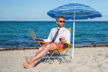 Sea Beach 'te serbest çalışan bir adam. Yazın iş adamı. Çevrimiçi serbest çalışma. Dizüstü bilgisayarı olan bir iş adamı. İş adamı plajda dizüstü bilgisayar kullanır. Seyahat ve iş. Şirket seyahat politikası.