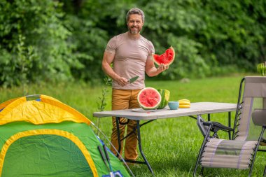 Adam kampı. Karpuz kampı pikniği. Lezzetli sağlıklı yiyecekler. Kampta taze yiyecek. Yaz kampında karpuzlu bir adam. Yaz günü doğa yürüyüşçüsü. Kamp ve doğa pikniği. Organik yiyecekler.