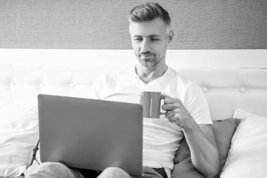 smiling mature man using computer in bed with coffee.