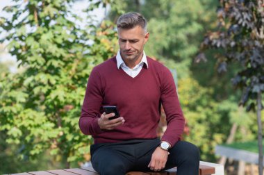 Olgun adam cep telefonu kullanır. İş adamı dışarıdaki telefonu kullanır. Oturan olgun bir iş adamı. Sonbahar tarzı. Sonbahar parkındaki iş adamı. Kafkasyalı bir adamın günlük bir tarzı vardır. Bir mesaj gönderiliyor.