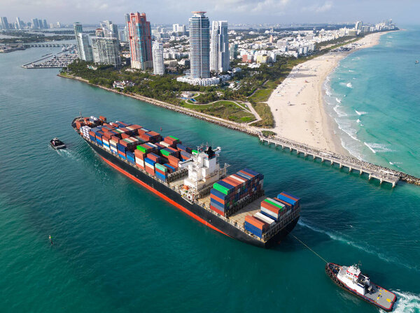 Грузовой корабль с контейнером в Майами, антенна. Грузовой контейнер на грузовом корабле. Грузовое судно покидает порт. Контейнерный корабль. Экспорт и импорт. Грузовой бизнес. Порт Майами. Океанское судно.
