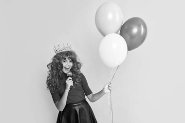 positive child in crown with microphone and party balloon on yellow background.