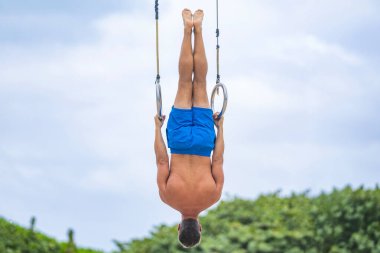 Spor yüzüklerinde asılı kaslı jimnastikçi. Güçlü bir atlet. Adam yanaşıyor. Arka görüşlü kaslı bir sporcu. Spor barfiks egzersizi. Spor eğitimi. Jimnastik yüzüklü bir sporcu. Baş aşağı asılı..