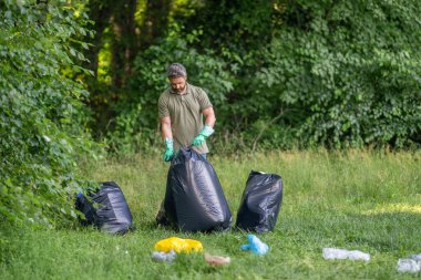 Ormanda çöp toplama. Ekoloji konsepti. Çöpleri toplayan gönüllü. Çöp torbasıyla lastik eldivenli bir adam çöpü temizliyor. Adam plastik çöp topluyor. Çevre gönüllüsü. Plastik atık sorunu.