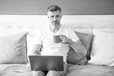 mature man smile using computer in bed with coffee.