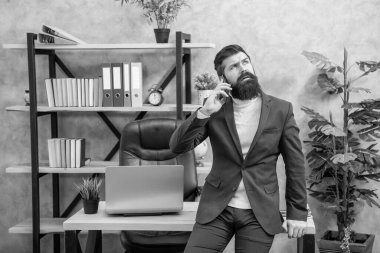 Takım elbiseli ciddi bir çalışan cep telefonuyla konuşuyor. Ofis masasında oturuyor, iletişim kuruyor..