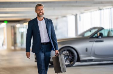 İş adamı evrak çantasını tutar. Başarılı bir anlaşmadan sonra takım elbiseli mutlu bir işadamı. Şirket işi. Takım elbiseli zengin iş adamı. İş portresi. Başarılı bir iş adamı park yerinde evrak çantasıyla yürüyor..