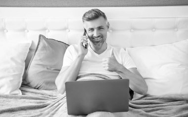 Olgun bir adam telefonda konuşuyor ve laptopla yatakta çalışıyor. başparmak yukarı.