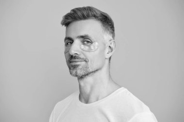 Göz altı kırışıklık önleyici krem sürmüş olgun bir adam. Yüz güzelliği cilt bakımı. Kırışıklık önleyici göz bandı olan adam mavi üzerine izole edilmiş. Yenilenme etkisi. Kozmetoloji. Siyah halkalarla savaş.