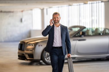 Takım elbiseli olgun bir iş adamı iş görüşmesi yapıyor. Şirket işi. Takım elbiseli zengin iş adamı. İş portresi. Başarılı bir iş adamı park yerinde evrak çantasıyla yürüyor. İş adamı evrak çantasını tutar.