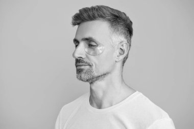 Kırışıklık önleyici göz bandı olan adam mavi üzerine izole edilmiş. Yenilenme etkisi. Kozmetoloji. Göz altı kırışıklık önleyici krem sürmüş olgun bir adam. Yüz güzelliği cilt bakımı. Şişkinliği azalt.