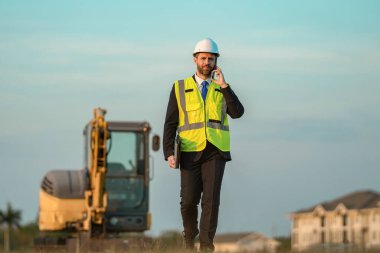 Bina sorumlusunun telefonu var. Hardhat 'ın baş mühendisi telefon kullanıyor. İş adamı telefonda konuşur. İnşaat mühendisi. Denetmen iş adamı. Şantiyedeki adam. İş projesi iletişimi.