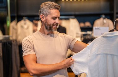 Tişört seçen olgun bir adam. Tasarımcı kıyafeti. Lüks mağazada günlük tarz erkek giyim. Şık bir adam için lüks butik alışverişi. Erkek giyim mağazası. Erkek giyim mağazasında şık bir adam. Kaliteli kumaş.