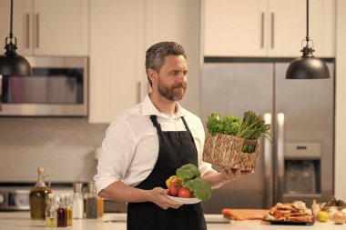 İspanyol adam eve sağlıklı yemekler yapıyor. Önlükle. Mutfaktaki yemek menüsü için malzeme seçiyor..