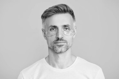 Kozmetoloji. Göz altı kırışıklık önleyici krem sürmüş olgun bir adam. Yüz güzelliği cilt bakımı. Kırışıklık önleyici göz bandı olan adam mavi üzerine izole edilmiş. Yenilenme etkisi. Yeterince uyu..