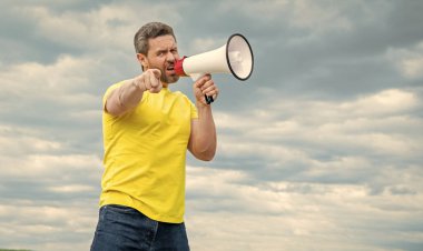 man in yellow shirt shout in megaphone on sky background. pointing finger.