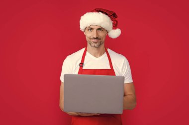 Noel Baba şapkalı olgun bir adam ve kırmızı önlüklü dizüstü bilgisayar..