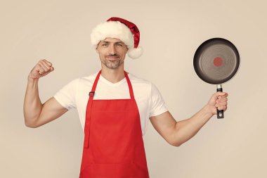 Kırmızı Noel Baba şapkalı ve önlüklü güçlü olgun adam kızartma tavasını tut..