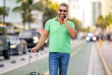 Yaz gezisini ayarlayan adam. İspanyol turist gezginleri kitabı internette. Seyahat çantalı bir turist. Gezgin telefon kullanıyor. Turist tatili rezervasyonu. Yaz gezisi için seyahat valizi. İnternetten rezervasyon.