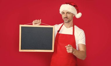 Yeni yıl şapkalı ve önlüklü neşeli olgun bir adam fotokopi alanı olan karatahtayı tutuyor. işaret parmağı.