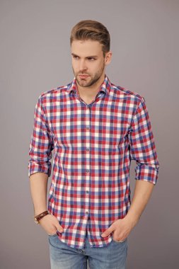 Kareli gömlek giyen milenyum adamı. Milenyum erkeğinin gündelik tarzı. Sıradan bir tişörtle genç bir adamın portresi. Tıraşsız adamın şık bir portresi. Gri yolda izole edilmiş bir adam. Damalı biçim.