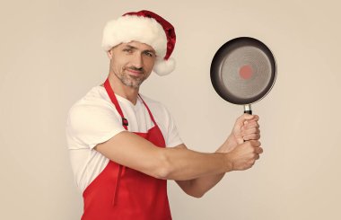 Kırmızı Noel Baba şapkalı ve önlüklü neşeli olgun adam kızartma tavasını tut..