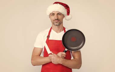 Kırmızı Noel Baba şapkalı ve önlüklü neşeli olgun adam kızartma tavası ve mutfak bıçağı tutuyor..