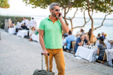 Yaz gezisi için seyahat valizi. Yaz gezisini ayarlayan adam. İspanyol turist seyahat kitabı internette. Seyahat çantalı bir turist. Gezgin telefon kullanıyor. Turist tatili rezervasyonu. Otel rezervasyonu.