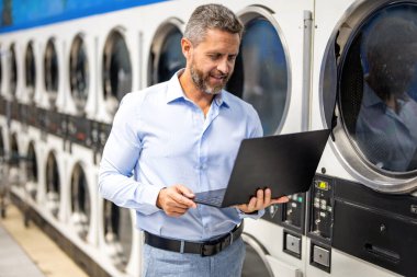 Çamaşır ve iş yükleme işinde çok çalışan var. Çamaşır ve teslim tarihleriyle uğraşan yorgun bir işçi. Yoğun iş adamları için aşırı çamaşır yüklemesi. Yoğun bir program. Çamaşırhanede dizüstü bilgisayarda çalışan meşgul bir adam..