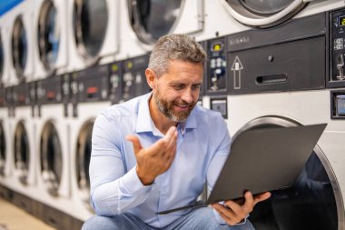 Yoğun iş adamları için aşırı çamaşır yüklemesi. Çamaşırhanede dizüstü bilgisayarda çalışan meşgul bir adam. Çamaşır ve iş yükleme işinde çok çalışan var. Çamaşır ve teslim tarihleriyle uğraşan yorgun bir işçi. Planlanan zamanda etkili planlama.