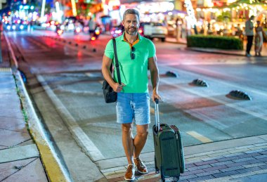 İnsan hafta sonu yurt dışına çıkıyor. Seyahat yaşam tarzı. Yaz şehrinde bir turist. Seyahat çantası. Amerika 'ya yaz gezisi. Miami 'de olgun bir turist. Bavullu sıradan giysili bir gezgin. Gece uçuşu.