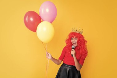 positive child in crown with microphone and party balloon on yellow background.