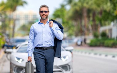 Başarılı bir iş adamı park yerinde evrak çantasıyla yürüyor. İş adamı evrak çantasını tutar. Takım elbiseli olgun iş adamı. Şirket işi. Takım elbiseli lüks iş adamı. İş portresi, kopyalama alanı.