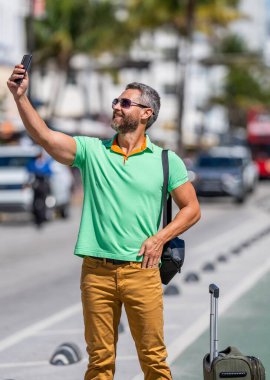 Adam yaz tatilinde selfie çekiyor. İspanyol turist gezginlerinin açık havada online video görüşmeleri var. Seyahat çantalı bir turist. Gezgin telefon kullanıyor. Turist tatili. Yaz gezisi için seyahat valizi..