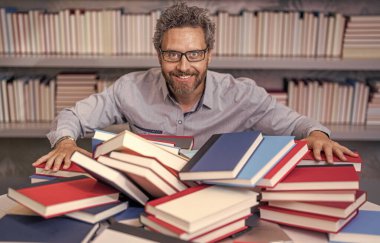 Okul kütüphanesinde dahi öğretmen. Kitap yığınında ev ödevi edebiyatı okumak dahice. Erkek öğretmen kitap okudu. Üniversitede okul eğitimi. Bilgi günü. Kitap kurdunun fazla çalışması. eğitim edebiyatı.