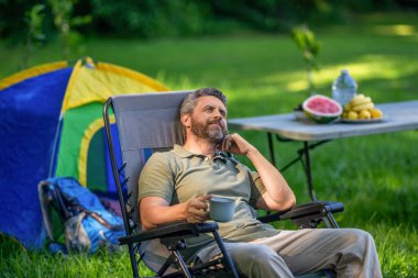 Adam ormanda sabah kampı kahvesi içiyor. Yürüyüşçü adam dışarıda rahatlar. Yaz kampı ve doğa tatili. Kamp yapmayı seven adam. Kamp macerası. Yürüyüş konsepti. Kamp çadırı. Kahve fincanı. Hayal kurmak.