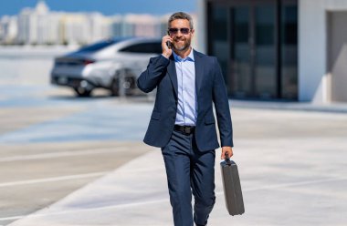 Takım elbiseli olgun bir iş adamı iş görüşmesi yapıyor. Şirket işi. Takım elbiseli zengin iş adamı. İş portresi. Başarılı bir iş adamı park yerinde evrak çantasıyla yürüyor. Şirket davası..