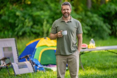 Adam ormanda sabah kampı kahvesi içiyor. Yürüyüşçü adam dışarıda rahatlar. Yaz kampı ve doğa tatili. Kamp yapmayı seven adam. Kamp macerası. Yürüyüş reklamı. Kamp çadırı. Kahve fincanı.