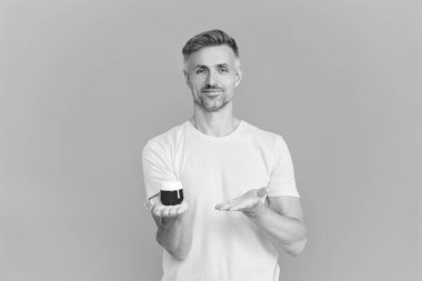 Mavi renkte yüz kremi gösteren bir adam var. Olgun adam cilt kremi kullanır. Günlük alışkanlık ve kişisel bakım. Cilt güzelliği. Erkek güzellik kozmetiği. Erkek kozmetik ürünü sunan adam. Nemlendirici uygula.