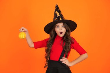 Cadılar Bayramı balkabağı elinde şaşkın cadı kız. Cadılar Bayramı cadısı, turuncu üzerine izole edilmiş. Cadılar Bayramı 'nda balkabağı. Cadıların Cadılar Bayramı kostümü. 31 Ekim balkabağı. Kostüm partisi.