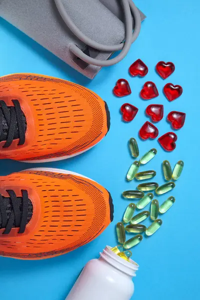 Yellow capsules of unsaturated fatty acids and red glass hearts, tonometer for measuring blood pressure, sports orange sneakers on a blue background. Vitamin supplements and exercise for heart health.