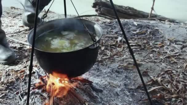 川岸の火災でカルドロンで川魚のスープを調理し スプーン付きの手は魚のスープをかき混ぜる — ストック動画