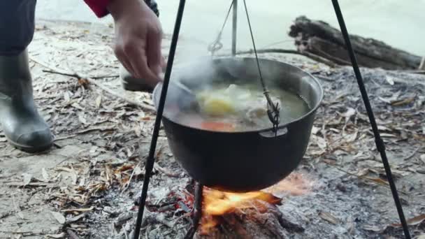 川岸の火災でカルドロンで川魚のスープを調理する スプーンを持つ漁師の手は 食欲をそそる魚のスープをかき混ぜる — ストック動画