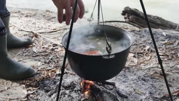 在河岸的火上 一个渔夫的手在锅里煮着河鱼汤 在鱼汤里加了干草 — 图库视频影像
